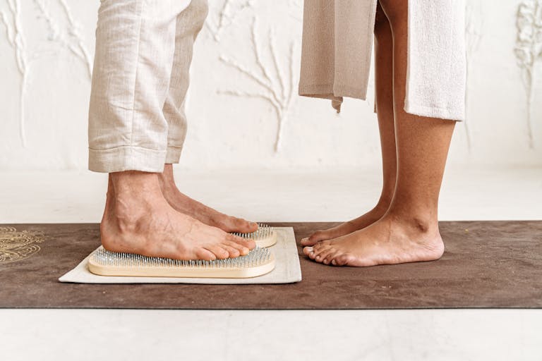 Two adults standing barefoot on an acupressure board, practicing reflexology indoors.